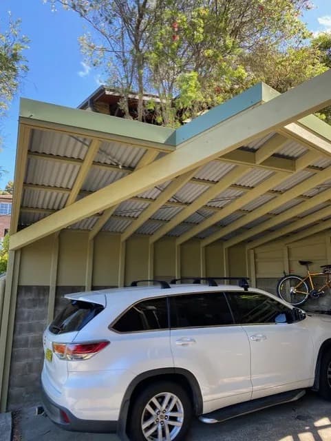 Timber-framed carport with painted posts and a corrugated roof, with a car parked beneath